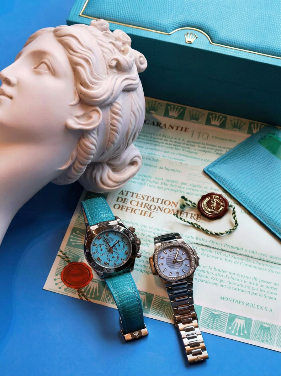 Two luxury Rolex watches resting on warranty certificates beside a marble bust and turquoise box