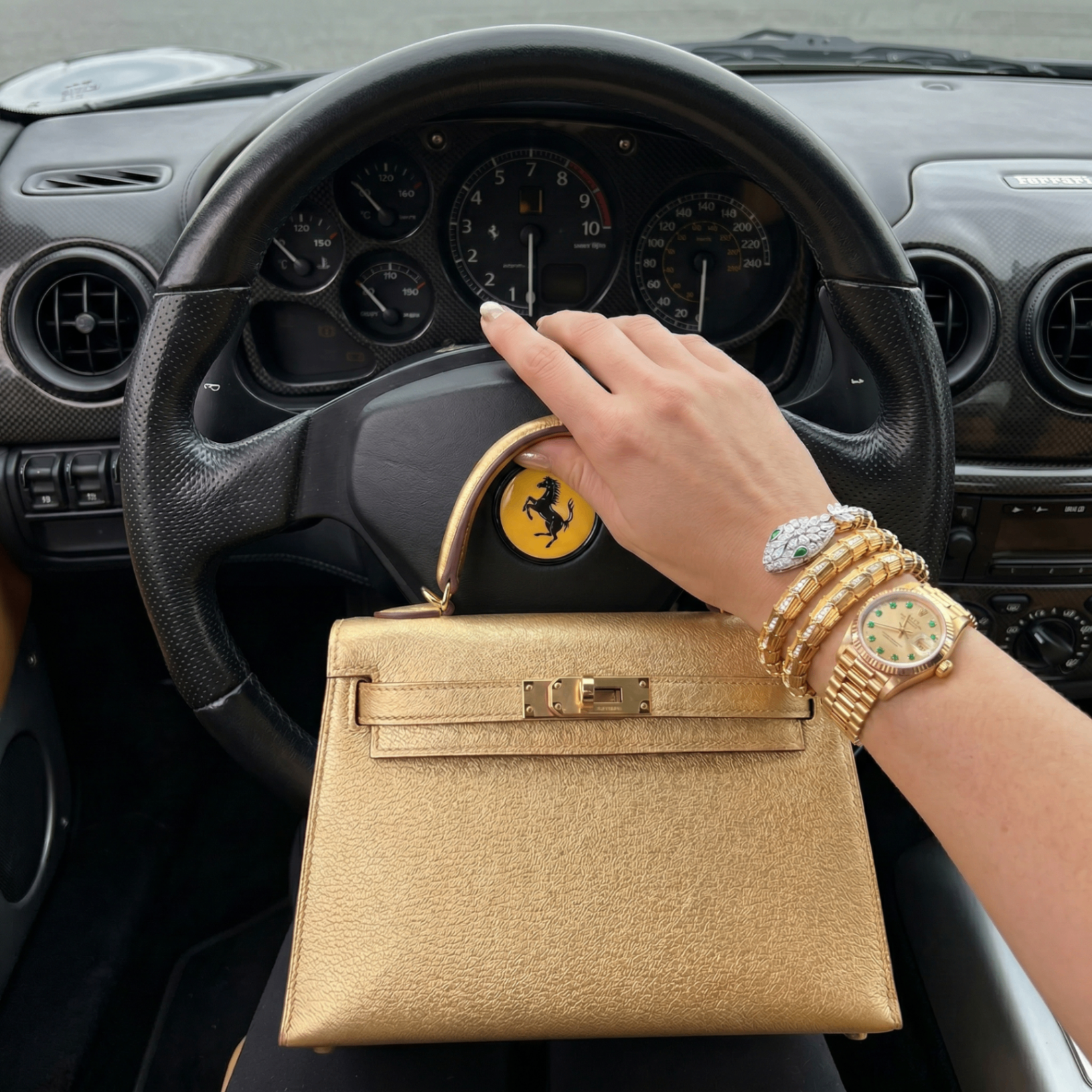 A hand on a Ferrari steering wheel holding a gold mini Kelly handbag with stacked bracelets